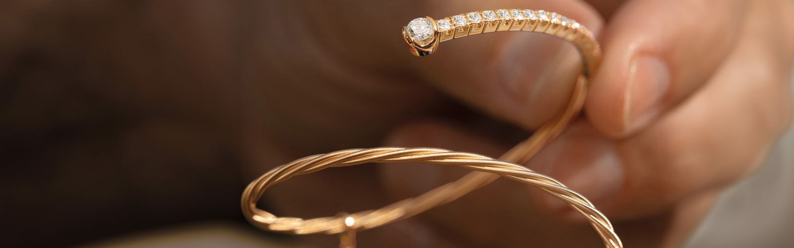 Eine Hand hält ein Wellendorff Armband mit funkelnden Brillanten an den Enden vor einem unscharfen Hintergrund. Es ist das 18-karätige federnde Gold von Wellendorff.